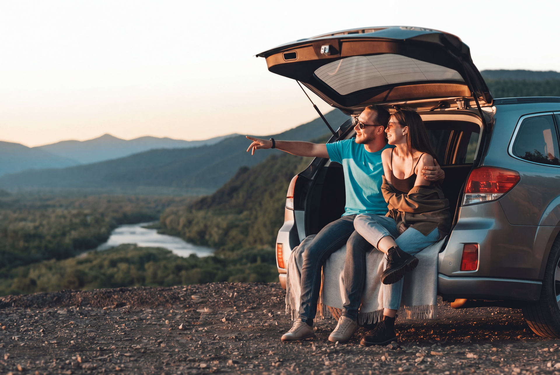 A couple sitting in the open trunk of an SUV looking out at mountains and a river at sunset