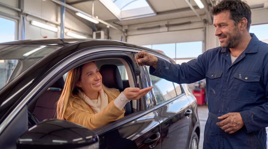 Mechanic handing car keys to a smiling customer seated in her car at an auto repair shop